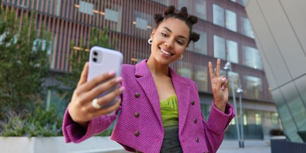 Attractive girl in fashionable outfit creats influence content shows peace sign at smartphone front camera poses for selfie smiles gladfully poses at urban place spends vacation at big city.