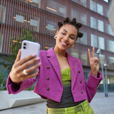 Attractive girl in fashionable outfit creats influence content shows peace sign at smartphone front camera poses for selfie smiles gladfully poses at urban place spends vacation at big city.